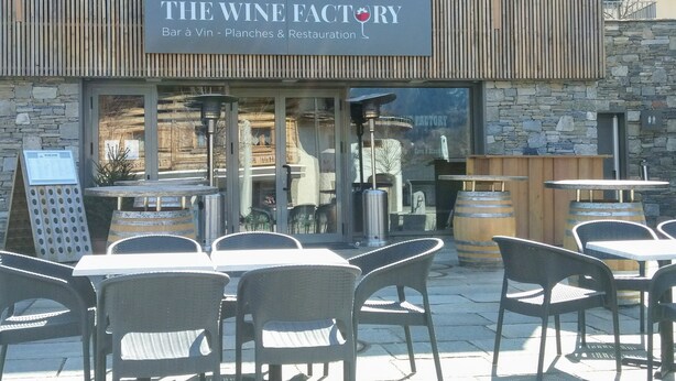 Un espace extérieur aménagé avec des tables et des chaises, situé devant un bâtiment portant l’inscription « THE WINE FACTORY Bar à Vin - Planches & Restaurant ».