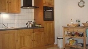 Une cuisine avec des meubles de rangement en bois, une hotte noire, un four en acier inoxydable et une petite table en bois sur laquelle se trouvent des bouteilles et une bouilloire.