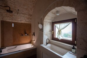 A bathroom with a stone wall, a wooden door, a window with a curtain, a sink, a mirror, and a shower.