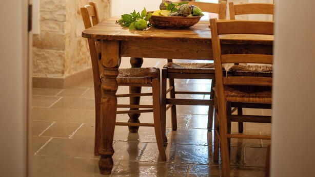 Un espace repas aux murs de pierre, avec une table et des chaises en bois, un saladier rempli de fruits et une vue dégagée sur l’extérieur, visible à travers une arche.