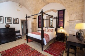 Un lit à baldaquin avec un mur en pierre en arrière-plan, une commode en bois, une table de chevet avec une lampe et une table à manger avec une chaise.