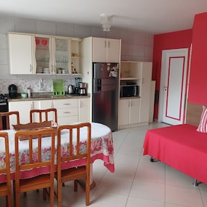 A compact kitchen with a refrigerator, microwave, and coffee maker. There is a dining table with chairs and a bed with a red bedspread. - Pousada Souza (Florianópolis)