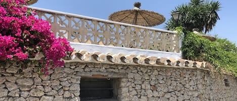 Eine Steinsäule mit einem dekorativen Balkon, einem Reetdachschirm und leuchtend rosa Bougainvilleenblüten.