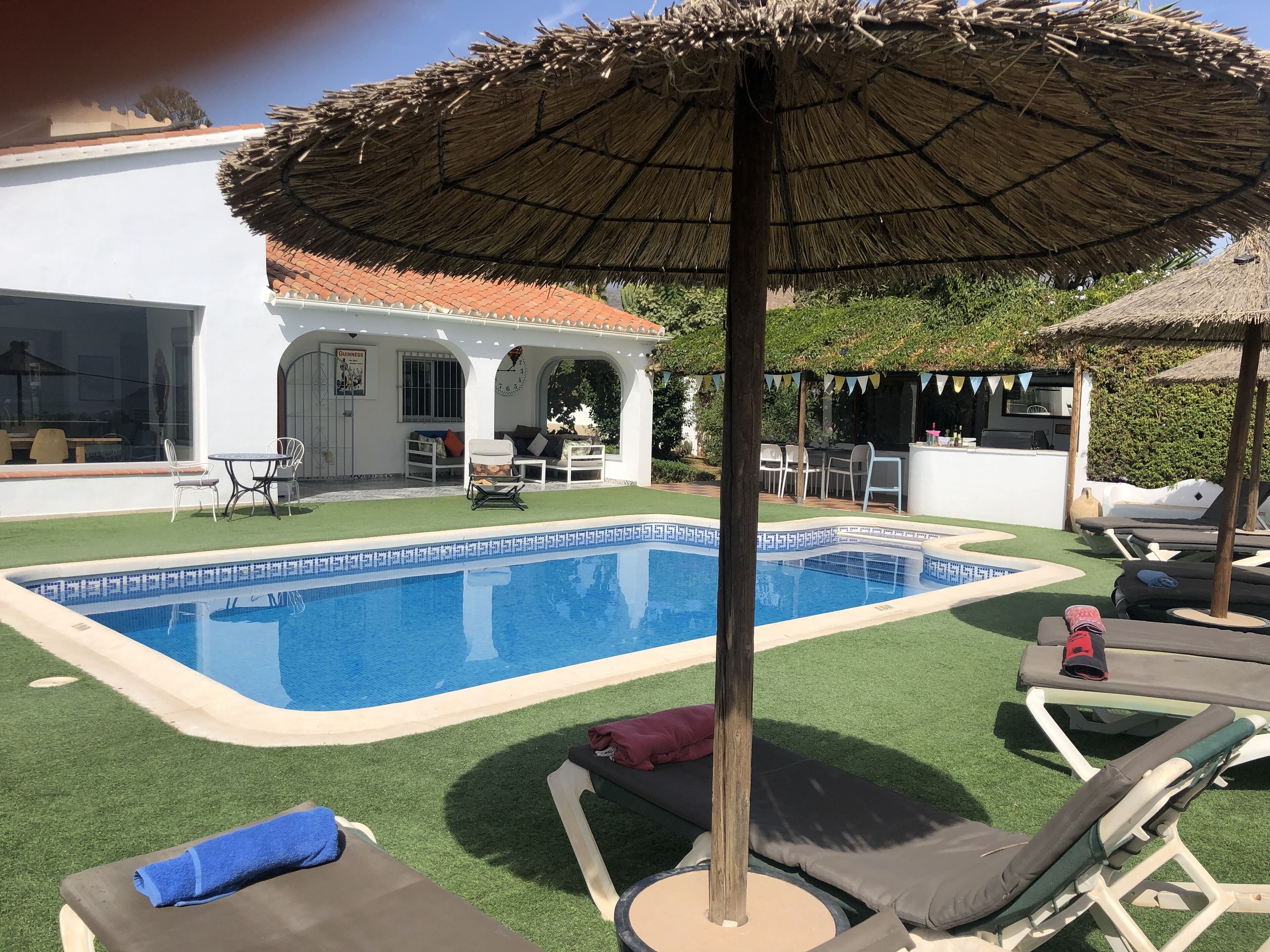 Una piscina con sillas de descanso y una sombrilla de paja, rodeada por un edificio blanco con techo de tejas.