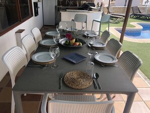 Een eettafel gedekt voor zes personen, met witte stoelen, een keuken op de achtergrond en een zwembad te zien door het raam.