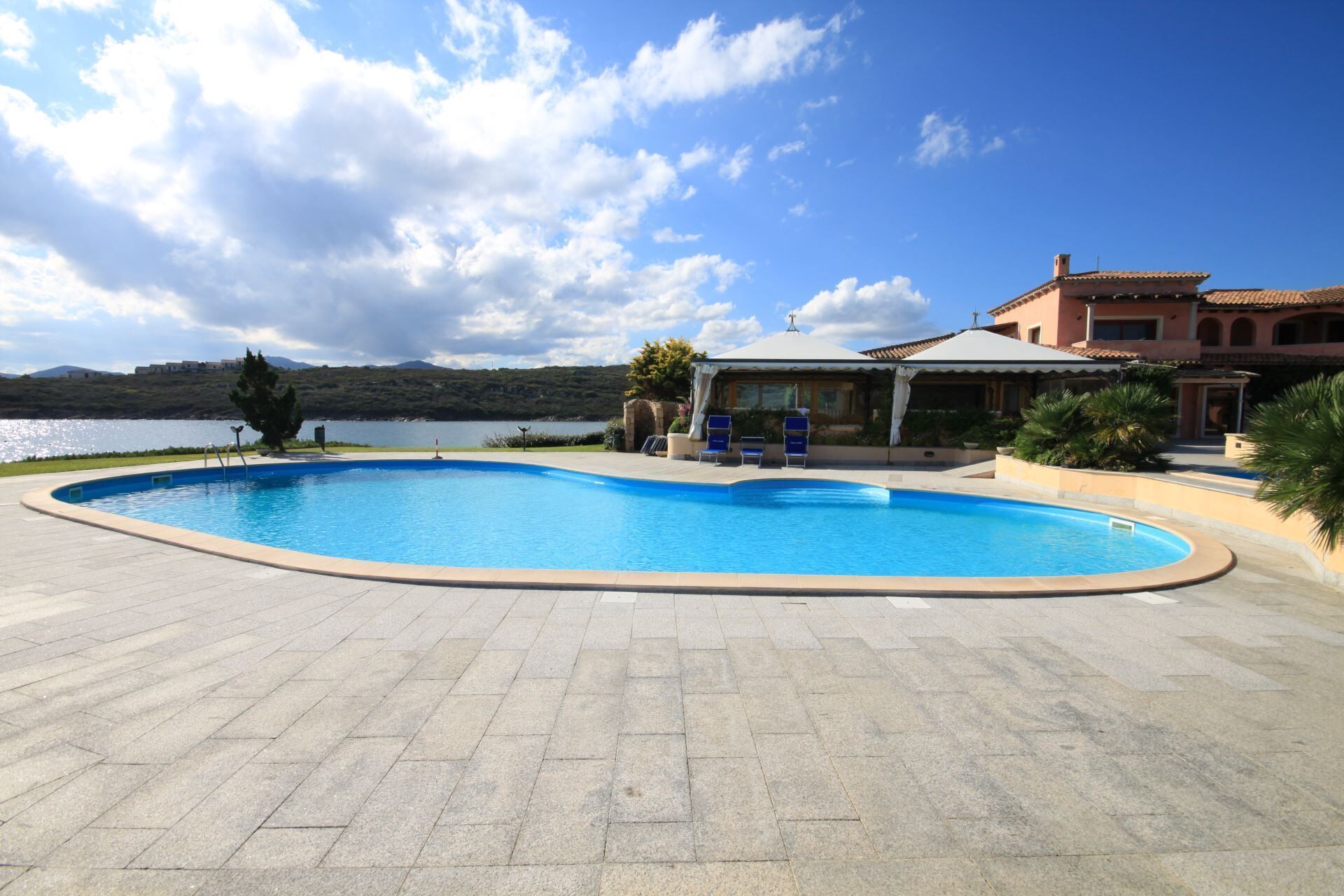 Una piscina de borde curvo, rodeada por una zona pavimentada. Hay sillas de descanso y una zona de estar cubierta. Al fondo se ve una masa de agua y colinas.