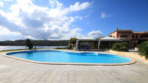A swimming pool with a curved edge, surrounded by a paved area. There are lounge chairs and a covered seating area. The background features a body of water and hills.