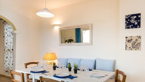 A dining area with a white table set for four, a blue patterned sofa, wooden chairs, a lamp, and wall art.