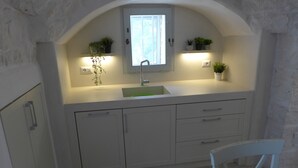 A kitchen with a white countertop, a sink, cabinets, and a window with plants on either side. - Beautifully Restored Trullo In Idyllic Location (Cisternino)