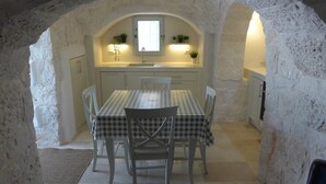 A stone-walled dining area with a checkered tablecloth, white chairs, a window above the sink, and potted plants on shelves. - Beautifully Restored Trullo In Idyllic Location (Cisternino)
