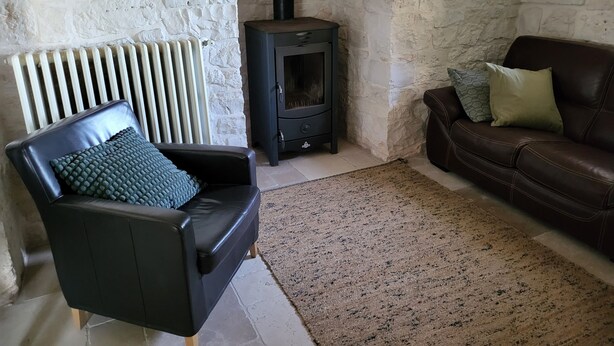 A cozy living room with a stone wall, a black leather sofa, a black armchair, a stone fireplace, and a radiator. - Beautifully Restored Trullo In Idyllic Location (Cisternino)