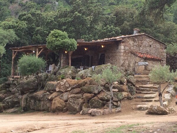 A rustic stone house with a tiled roof, surrounded by large rocks and trees.