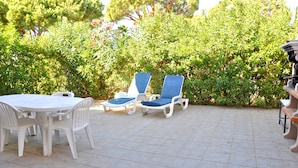 A patio with a round table and chairs, two blue lounge chairs, and a hedge in the background.