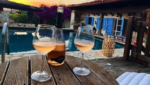 A wooden table with two wine glasses and a bottle of Akenta wine, set against a backdrop of a swimming pool and a sunset.