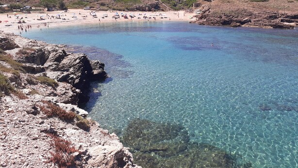 A beach with clear water, rocky shore, and a hilly background.
