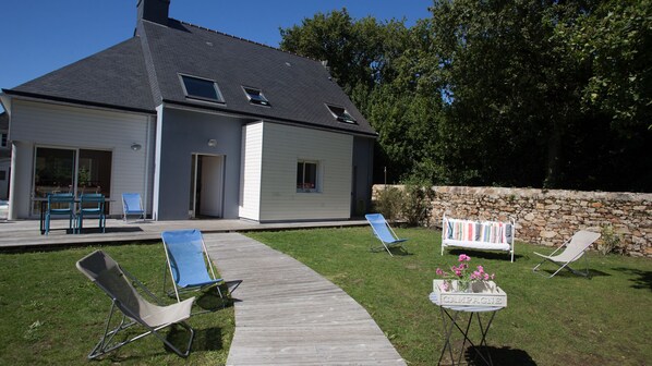 Une maison dotée d’un patio couvert, d’un mur en pierre et d’un jardin agrémenté de chaises longues et d’une petite table ornée de fleurs.