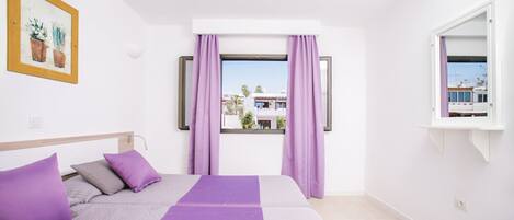 A bedroom with a bed, purple bedding, a window with curtains, a painting on the wall, and a view of buildings outside.