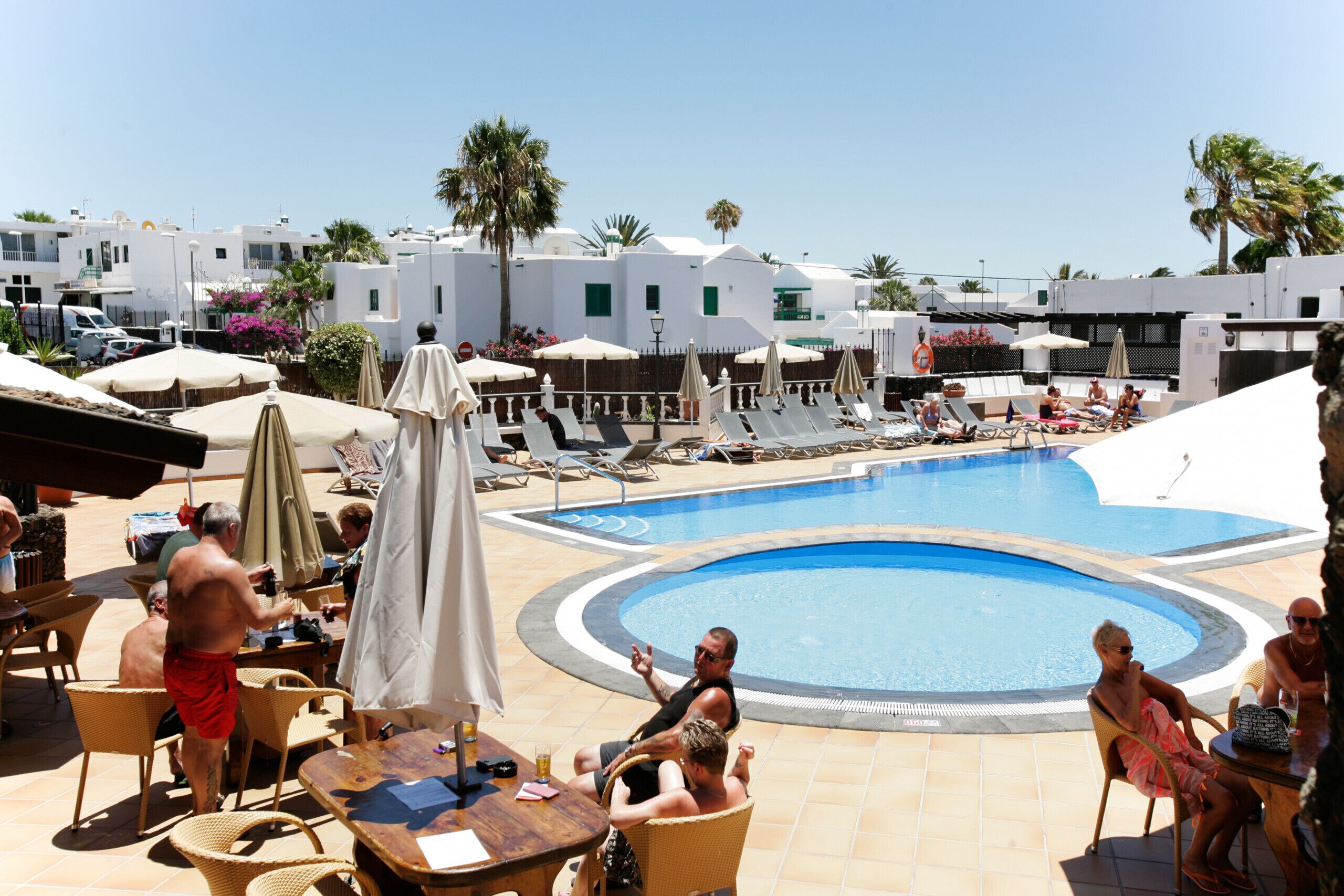 Un área de piscina con sillones de descanso y sombrillas, rodeada de edificios blancos y palmeras.