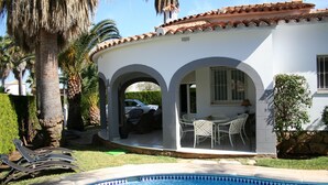 Zona de piscina con un edificio blanco, palmeras y un coche estacionado afuera.