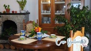 A dining area with a wooden table set for two, surrounded by potted plants and a wooden cabinet with glass doors containing dishes and glassware. - CASA BELLAVISTA ( “Bellavista” holiday home ) ……Where hospitality is at home (Furore)