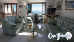 A living room with two green leather sofas, a glass coffee table with a vase of flowers, a television on a wooden cabinet, and a view of the sea through a large window. - CASA BELLAVISTA ( “Bellavista” holiday home ) ……Where hospitality is at home (Furore)