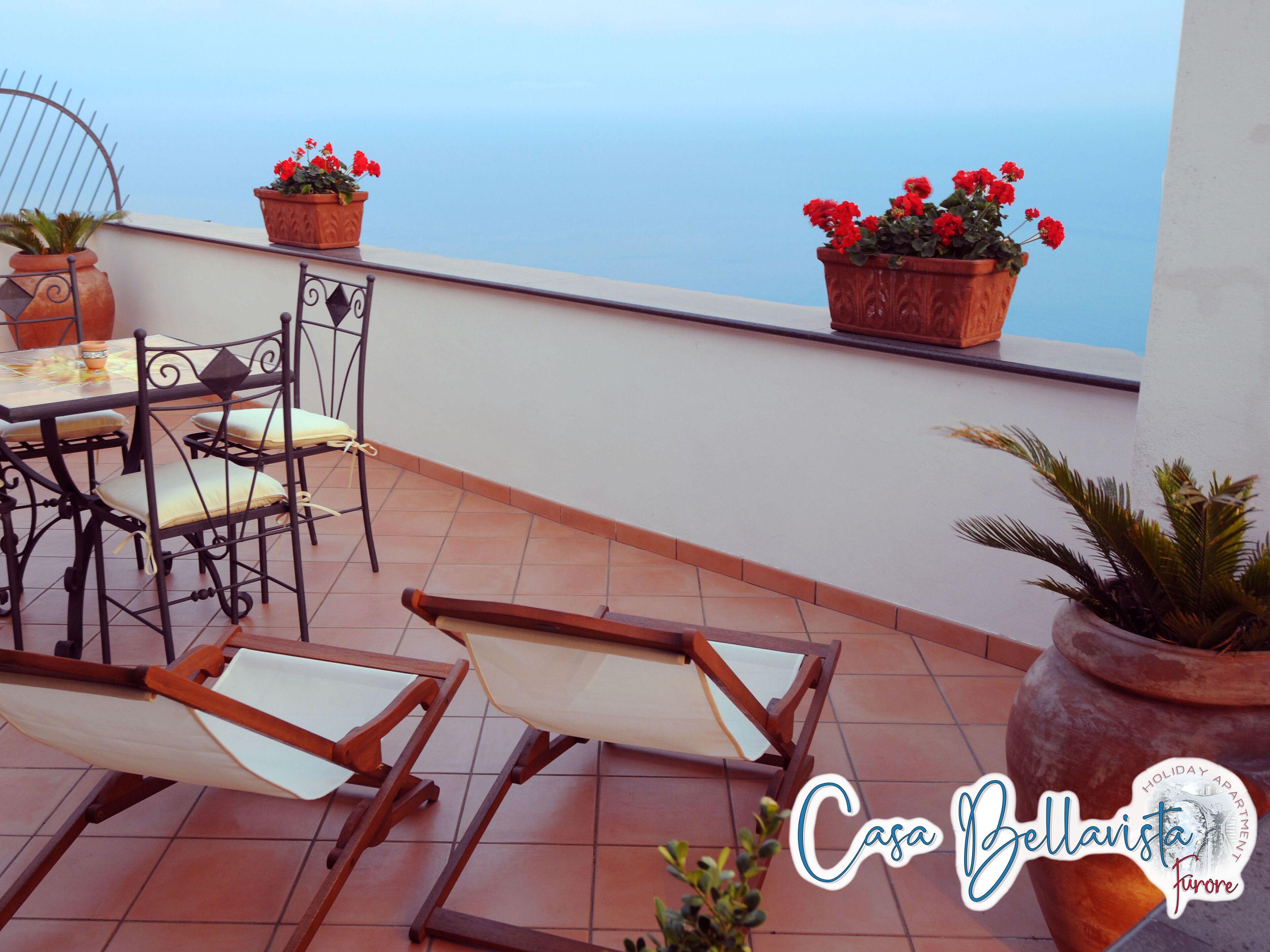 A terrace with a table and chairs, potted plants, and a view of the sea.