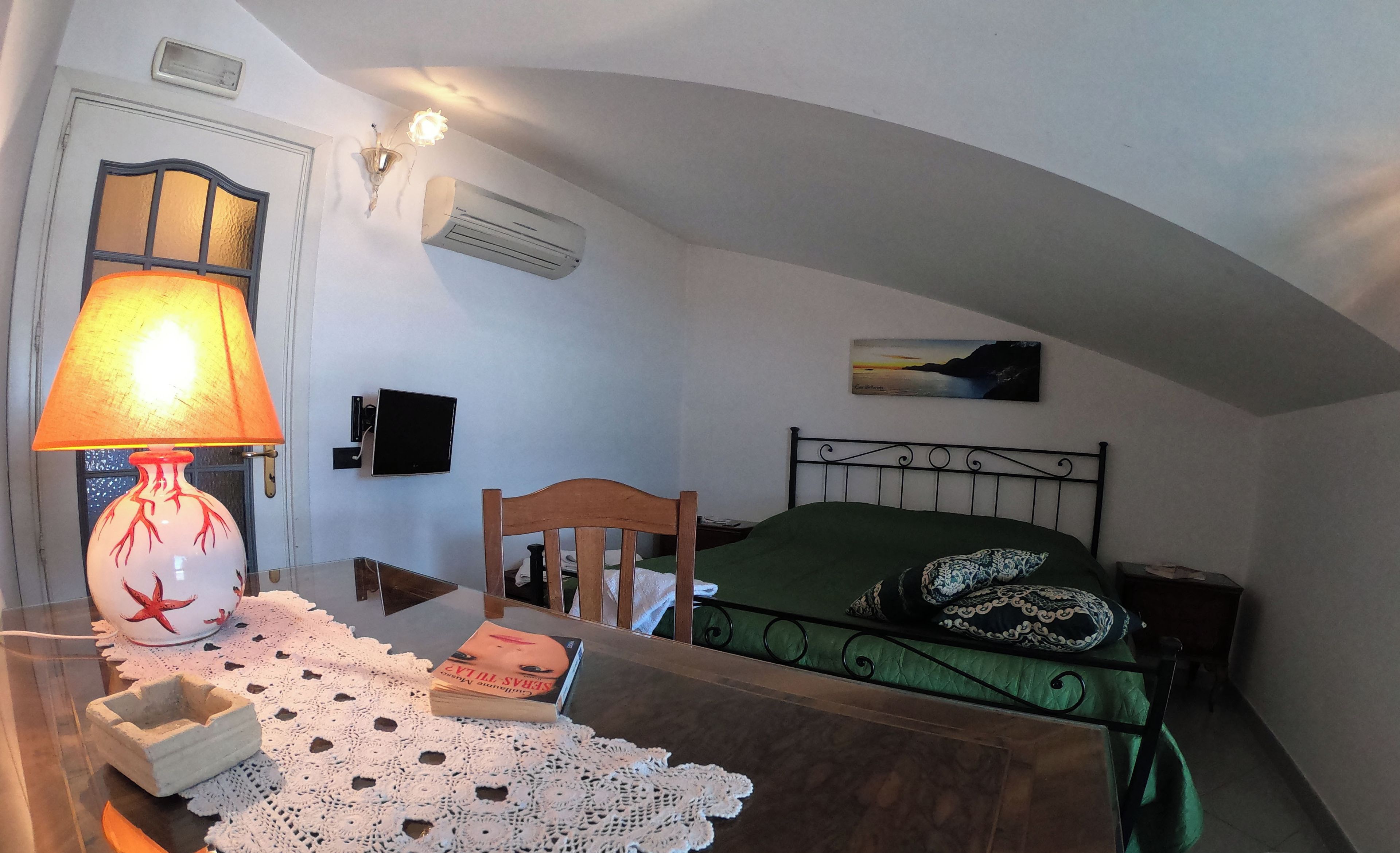 A bedroom with a wooden bed, a green bedspread, a white doily, a table lamp, a wall-mounted air conditioner, a television, and a painting of a sunset.