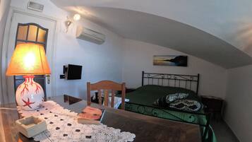 A bedroom with a wooden bed, a green bedspread, a white doily, a table lamp, a wall-mounted air conditioner, a television, and a painting of a sunset.