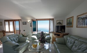 Un salon avec deux canapés en cuir vert, une table basse en verre ornée d’un vase de fleurs, un téléviseur posé sur un meuble en bois, et une vue sur la mer par le biais d’une grande fenêtre.