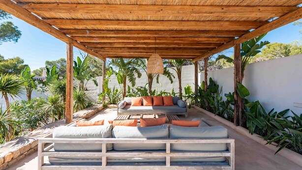 Un área de estar al aire libre con bancos de madera y cojines, rodeada de plantas tropicales y una pérgola de madera.