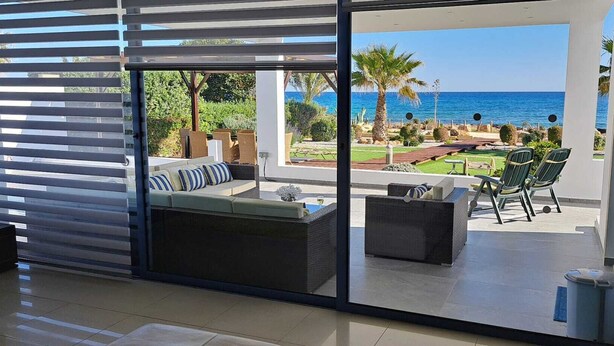 Una sala de estar moderna con un ventanal corredera que da a un patio con mobiliario exterior y vistas al mar.