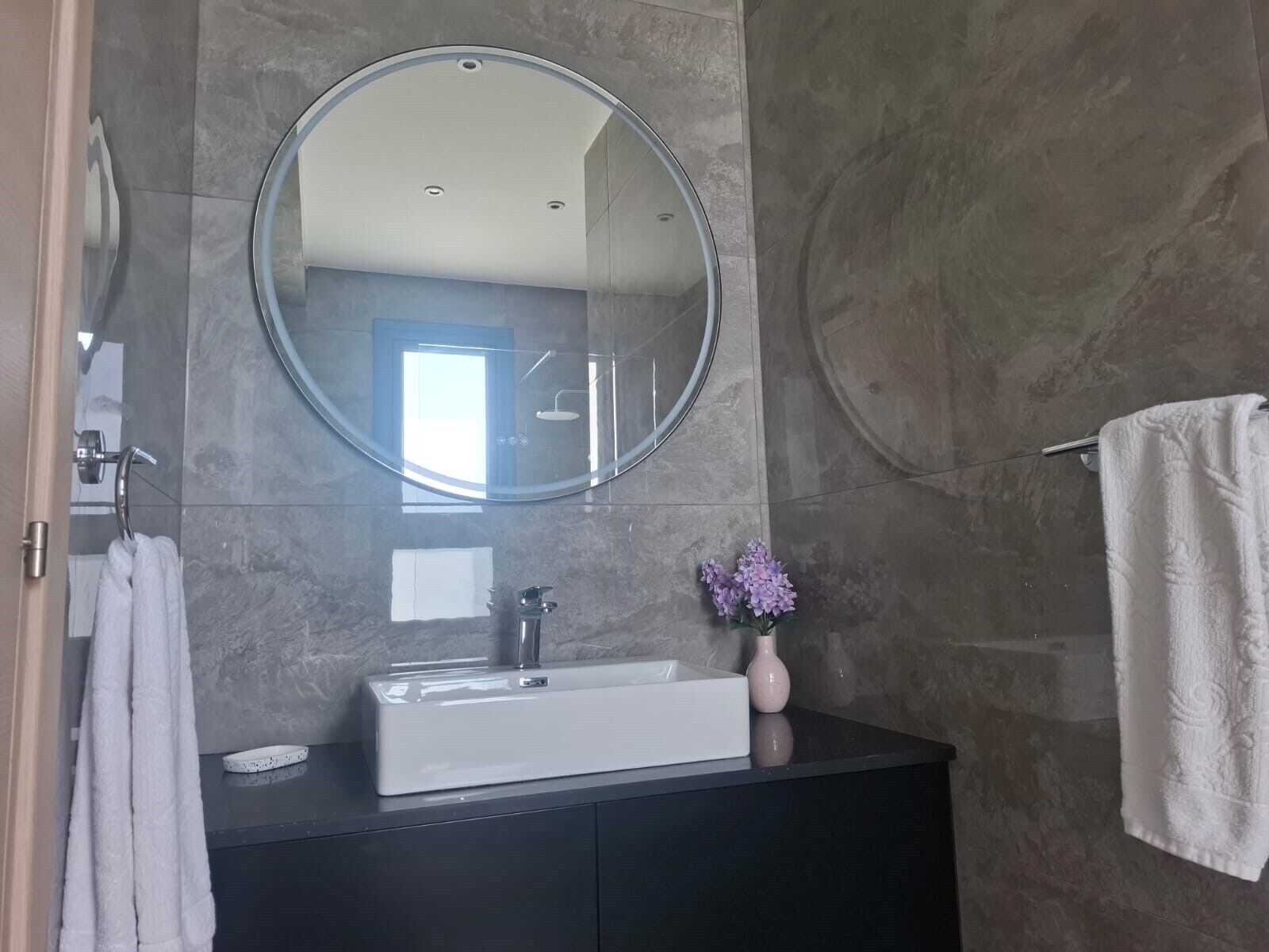 Un baño con un espejo grande, un lavabo blanco, un mueble de tocador negro y un perchero para toallas.