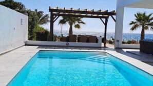 Una piscina rectangular con agua azul cristalina, rodeada por una terraza de baldosas blancas. Se observa una pérgola de madera con cojines y una vista al paisaje urbano y al océano de fondo.