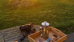 Une table en bois sur laquelle sont posés un appareil photo, un pot en métal, une tasse et un plateau avec une carafe et un verre, le tout sur fond de coucher de soleil au-dessus d’un champ d’herbe.