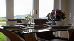 Une table à manger dressée avec des assiettes, des bols et des verres à vin, donnant sur l’extérieur grâce à de grandes fenêtres.