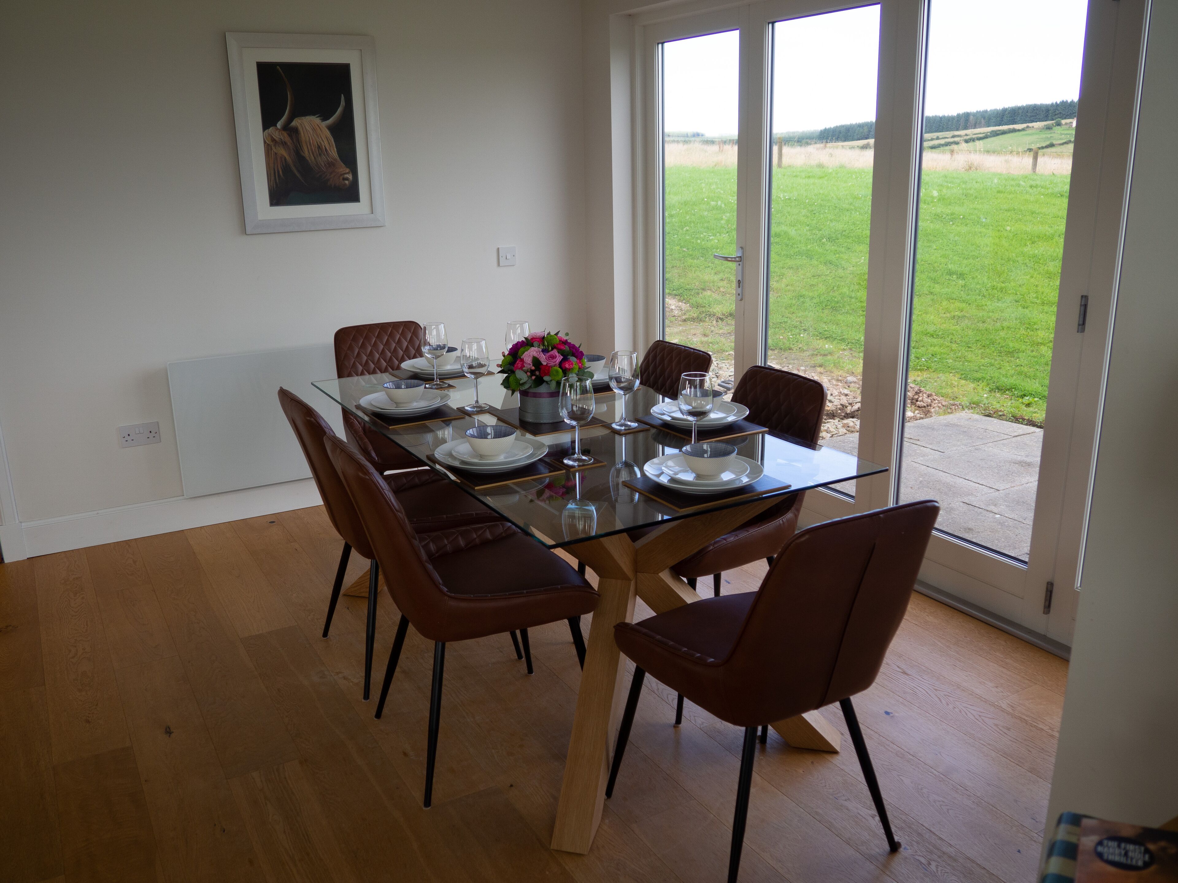 Un coin repas avec une table en verre, des chaises marron et une vue sur un champ verdoyant, visible à travers de grandes fenêtres.