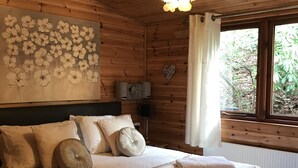 A wooden cabin bedroom with a bed, pillows, a wall art of flowers, a hanging light fixture, and a window with curtains. - Lake District Log Cabin (Troutbeck Bridge)