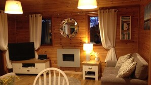 A cozy cabin interior with wooden walls, a television, a fireplace, a mirror, and a dining area. - Lake District Log Cabin (Troutbeck Bridge)