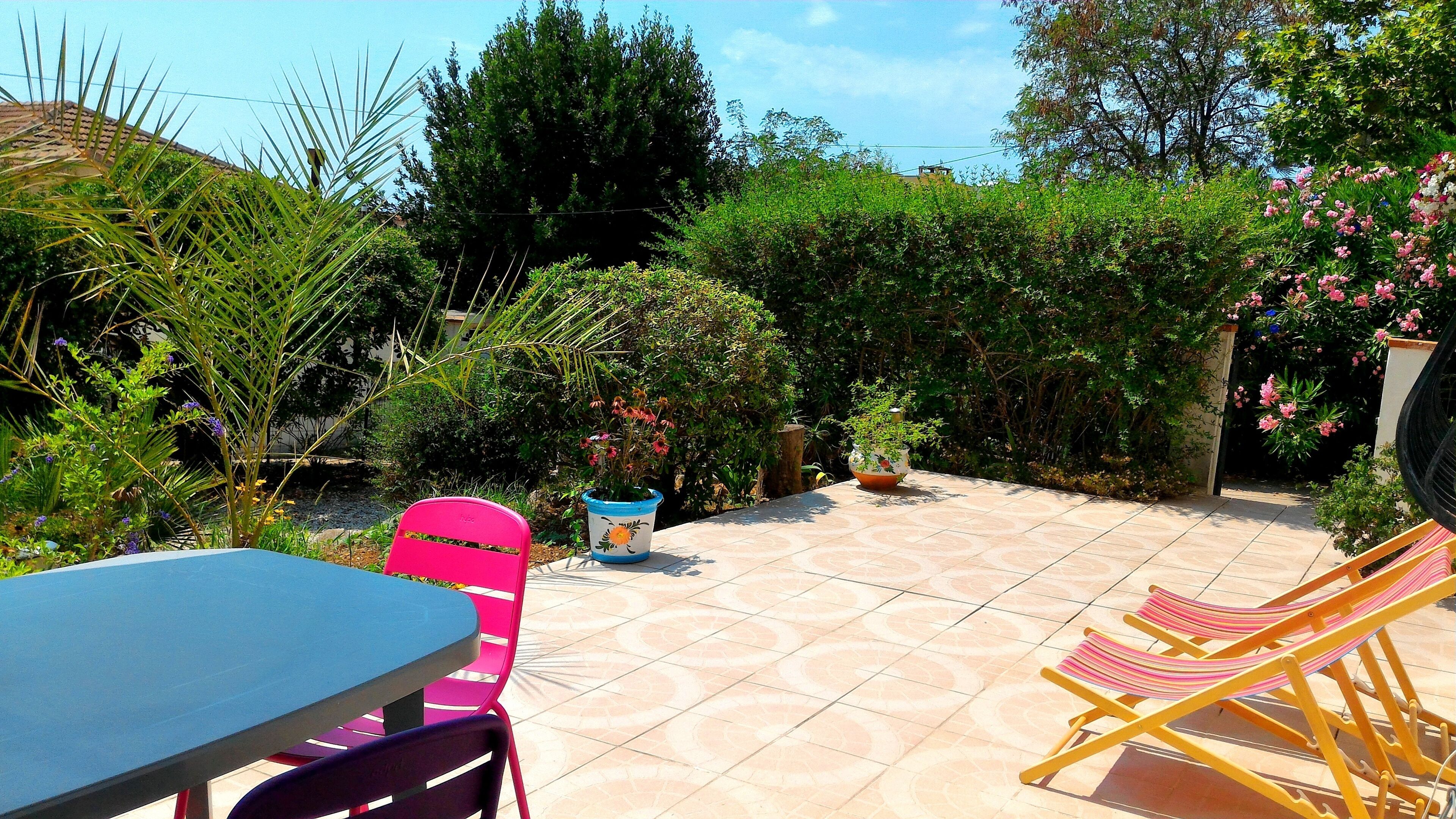 A patio with a table and chairs, surrounded by lush greenery and flowering plants.