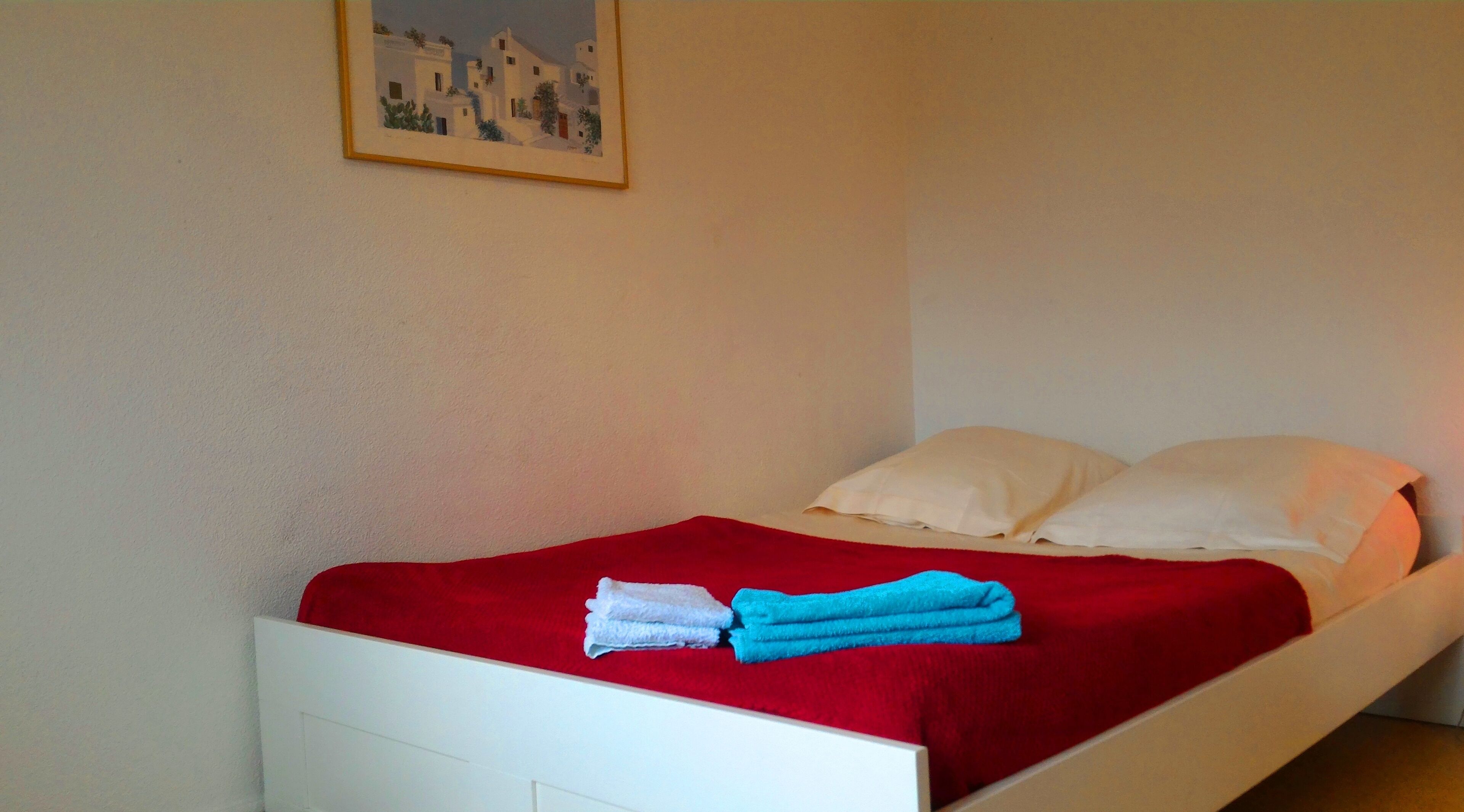 Ein Bett mit roter Decke, zwei Kissen und einem Bild an der Wand.