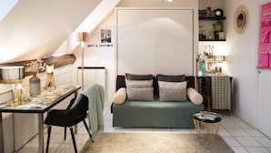 A cozy loft bedroom with a green sofa, a dining table with chairs, and a small round table with a lamp. - Studio With a Nice View of Paris - Luxury Building in the Heart of the Marais (Paris)