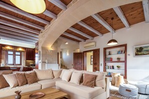 Un espacio habitable amplio con un sofá seccional beige grande, vigas de madera en el techo, una mesa de centro de madera y una estantería con libros y objetos decorativos.