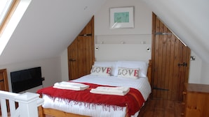 A bedroom with a sloped ceiling, wooden bed frame, white bedding, and a red throw blanket. - Rural Cotswold Cottage in grounds of beautiful Nell House (Hannington)