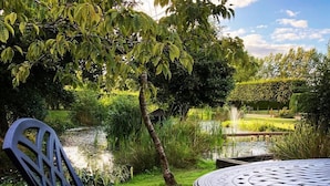A wooden deck with a metal table and chairs, surrounded by lush greenery and a pond. - Rural Cotswold Cottage in grounds of beautiful Nell House (Hannington)