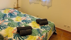 Ein Schlafzimmer mit einem Bett, das mit einer Bettdecke mit tropischem Blattmuster bezogen ist, zwei zusammengerollte Handtücher auf dem Bett, einer kleinen Holzhilfe mit einem dekorativen Gegenstand und einem Fenster mit durchsichtigen Vorhängen.
