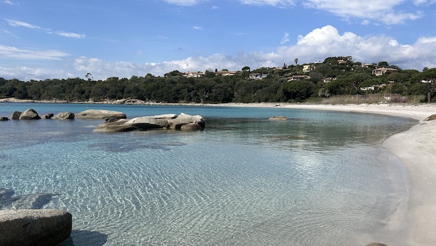Ein Strand mit klarem Wasser, großen Felsen und einer entfernten Ansicht von Häusern an einem Hügel.