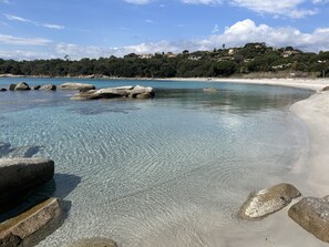Une plage à l’eau claire, bordée de gros rochers, d’où l’on aperçoit, au loin, des maisons sur une colline.