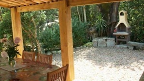 Eine Terrasse im Freien mit einem Holztisch und -stühlen, umgeben von Grünpflanzen und einem Grillplatz aus Stein.