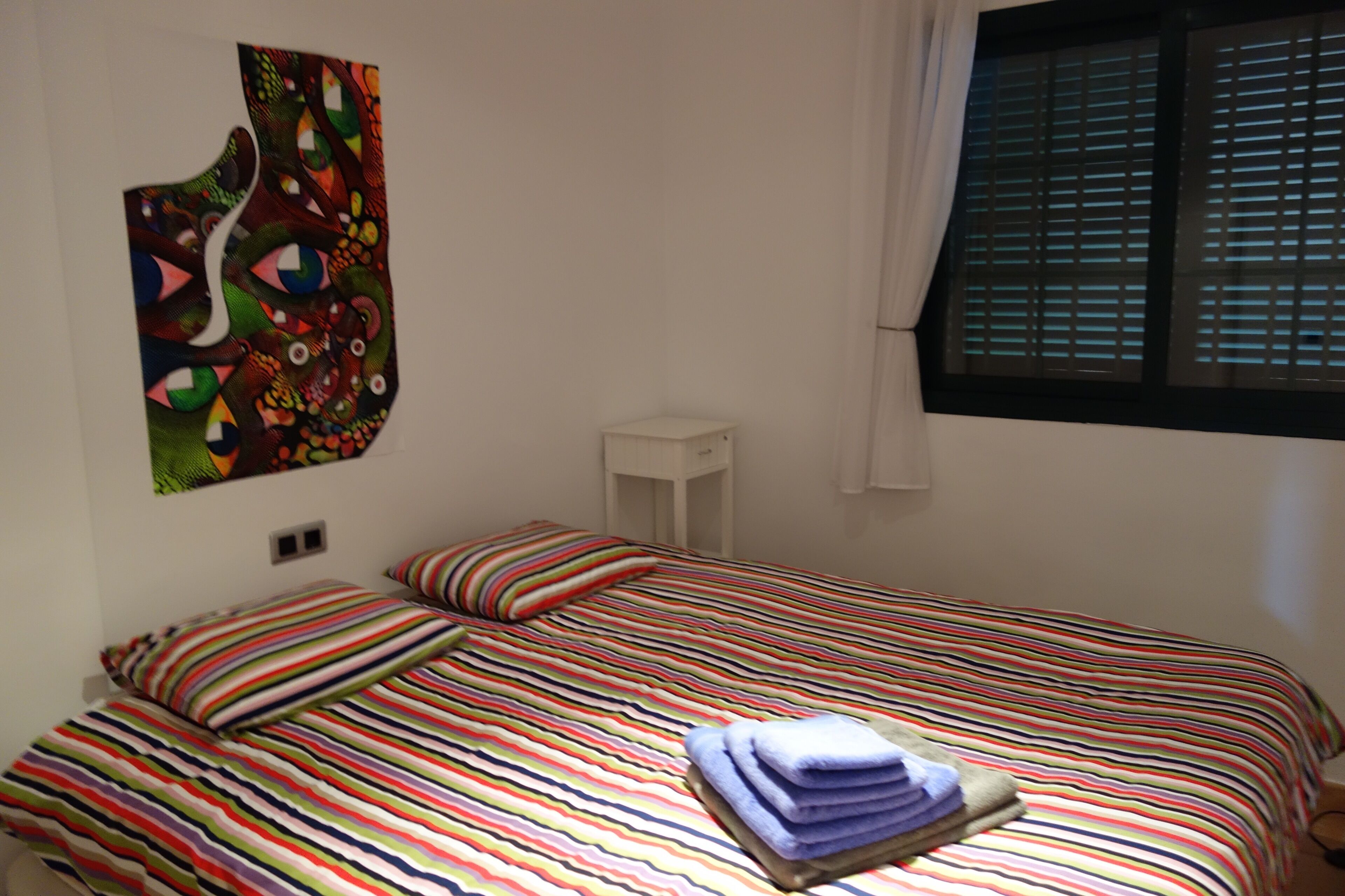 A bed with a striped bedspread, a colorful abstract wall hanging, and a window with blinds.
