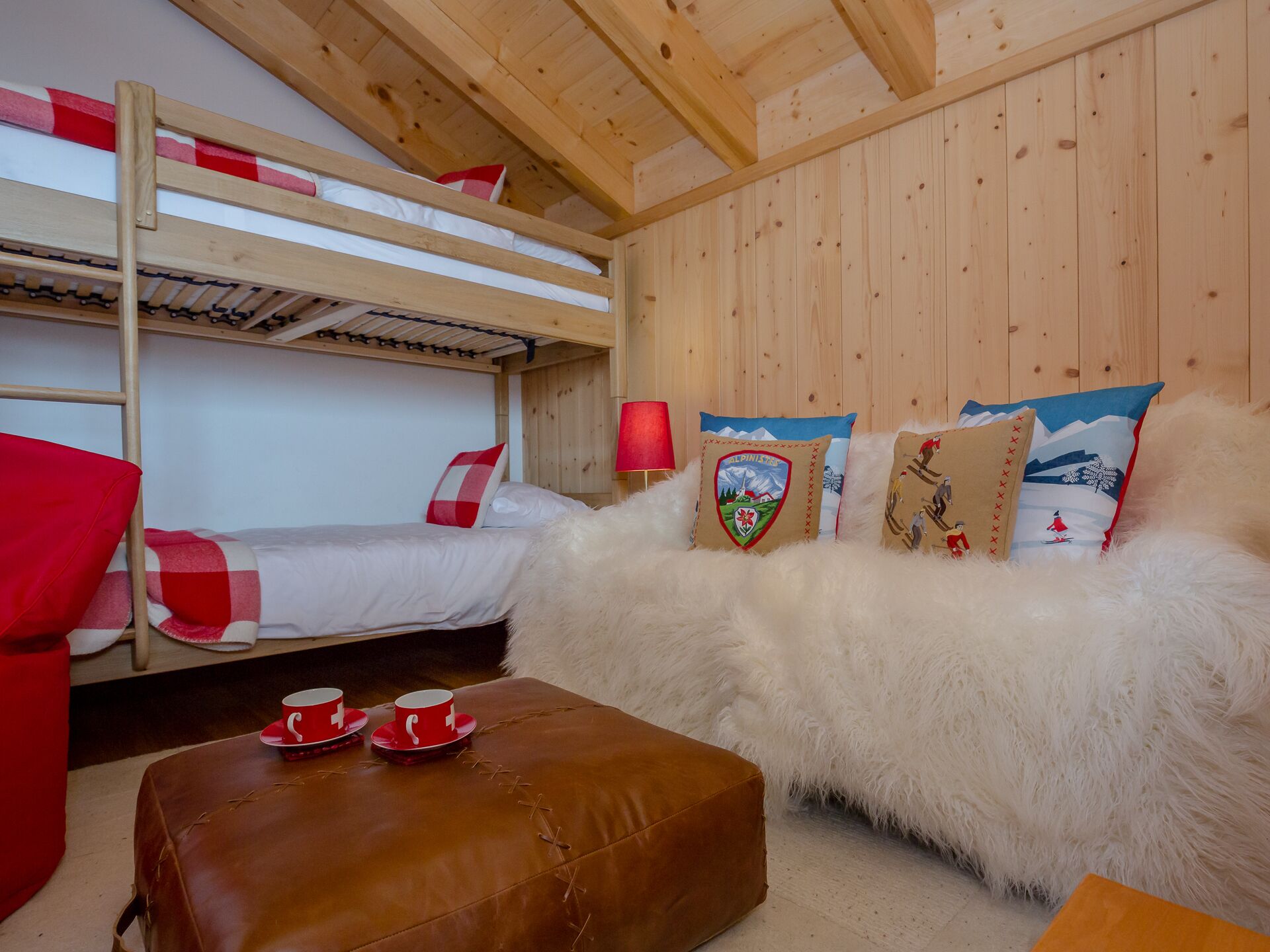 Een slaapkamer met een stapelbed, een houten plafond, een witte, pluizige bank en een bruin voetenbankje met rode bekers.